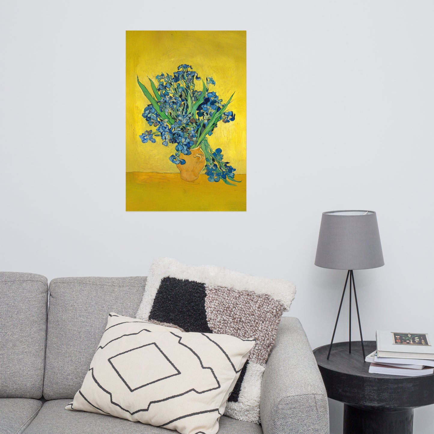 Van Gogh's 'Vase with Irises Against a Yellow Background' on a 24"x36" unframed poster print on a white wall above a gray sofa with throw pillows and side table with a lamp.