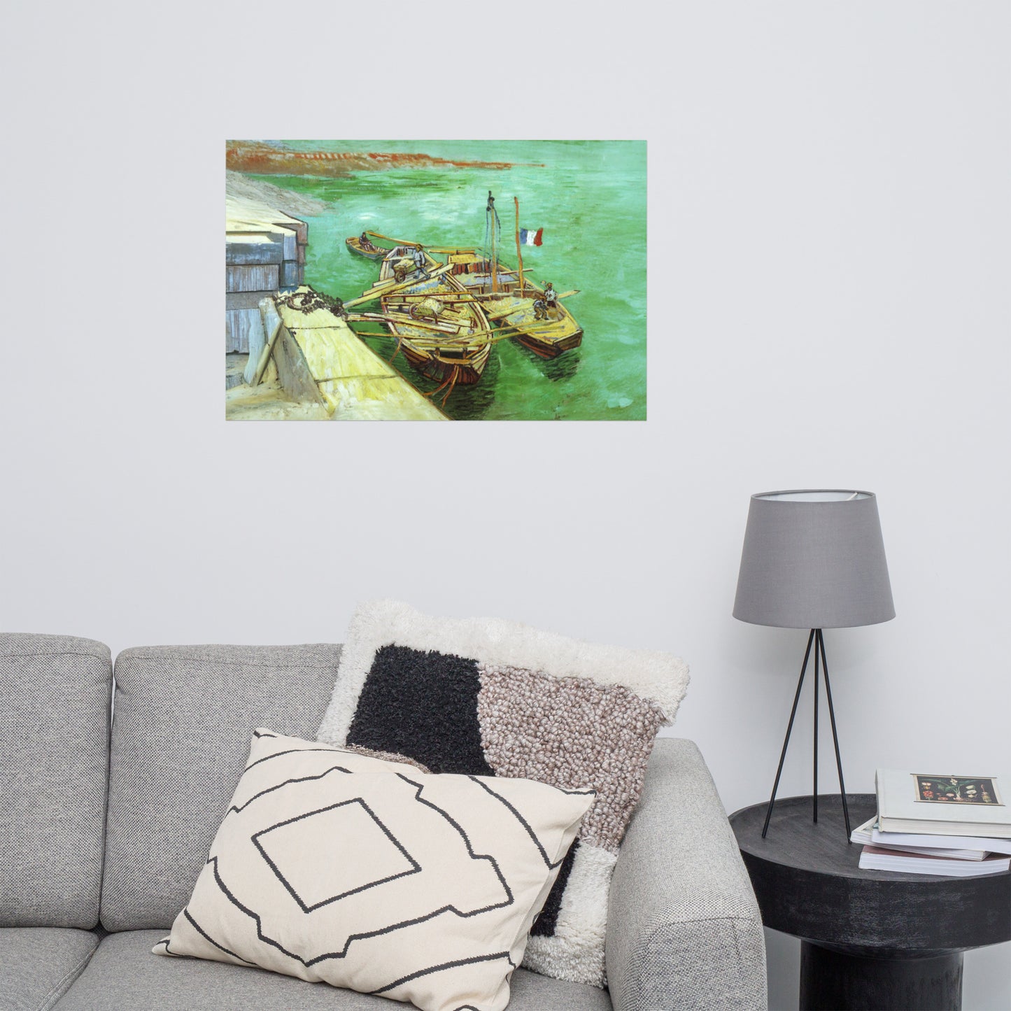 Van Gogh's 'Quay with Men Unloading Sand Barges' on a 24"x36" unframed poster print on a white wall above a gray sofa with throw pillows and side table with a lamp.