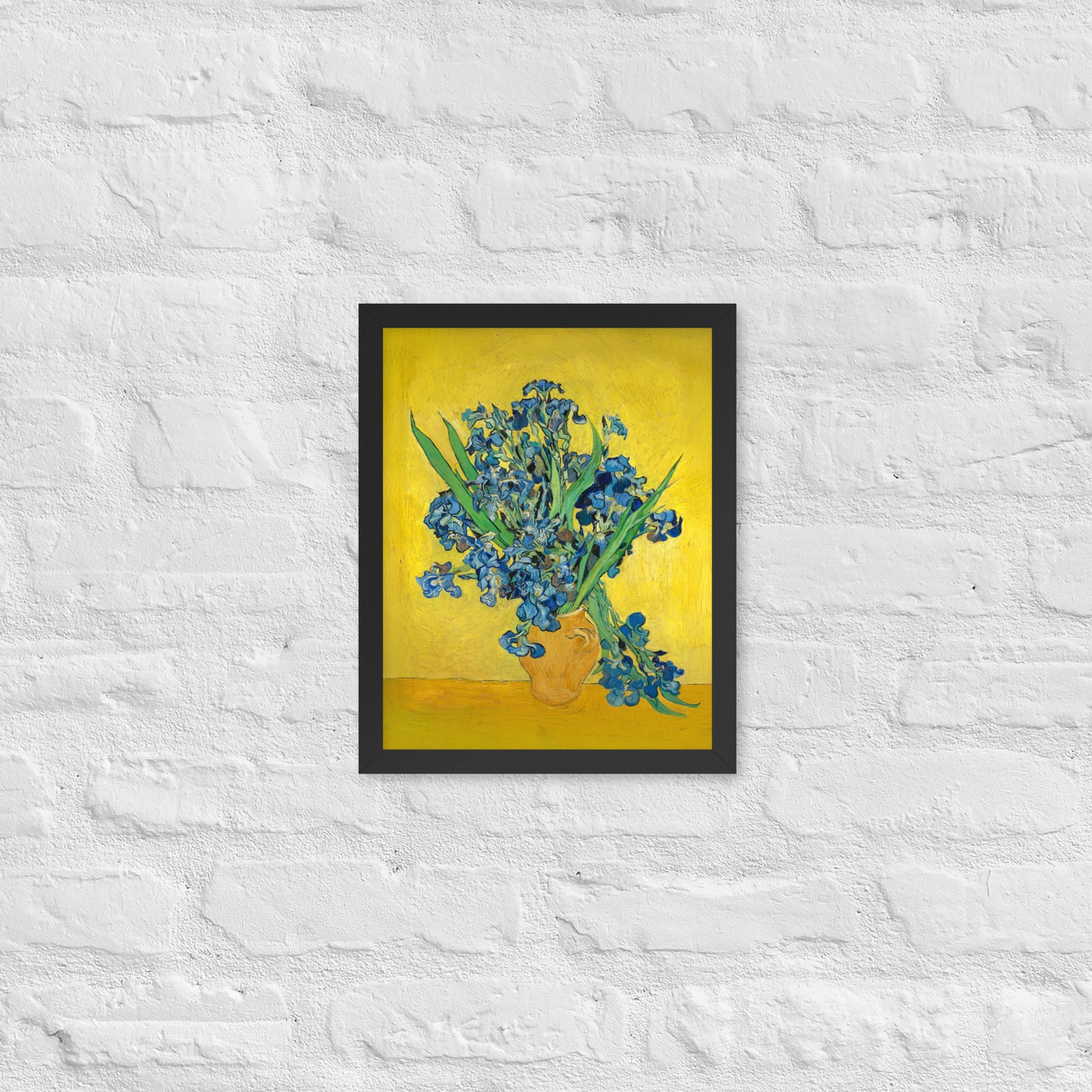 Van Gogh's 'Vase with Irises Against a Yellow Background' on a 11"x14" black framed poster print on a white brick wall.