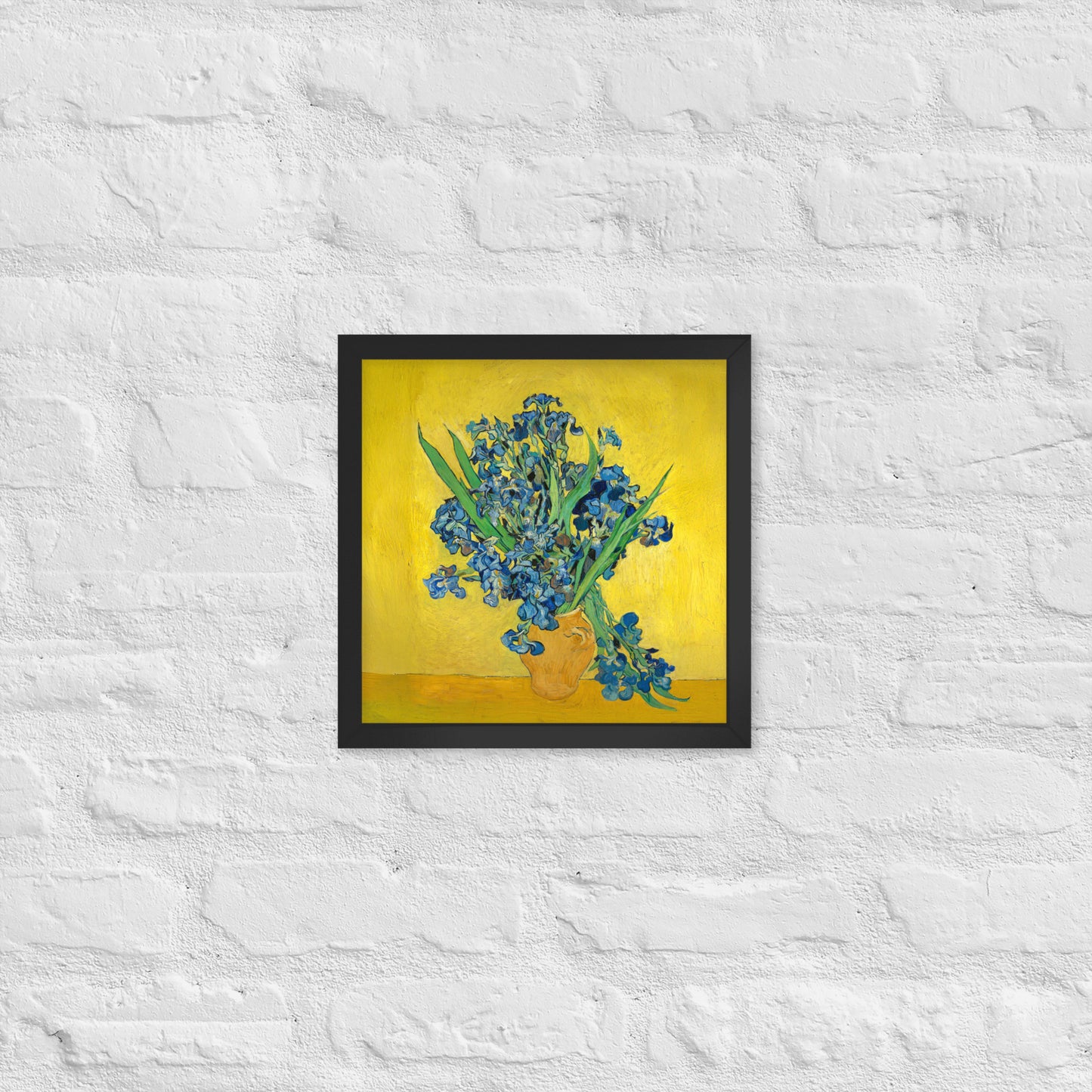 Van Gogh's 'Vase with Irises Against a Yellow Background' on a 12"x12" black framed poster print on a white brick wall.