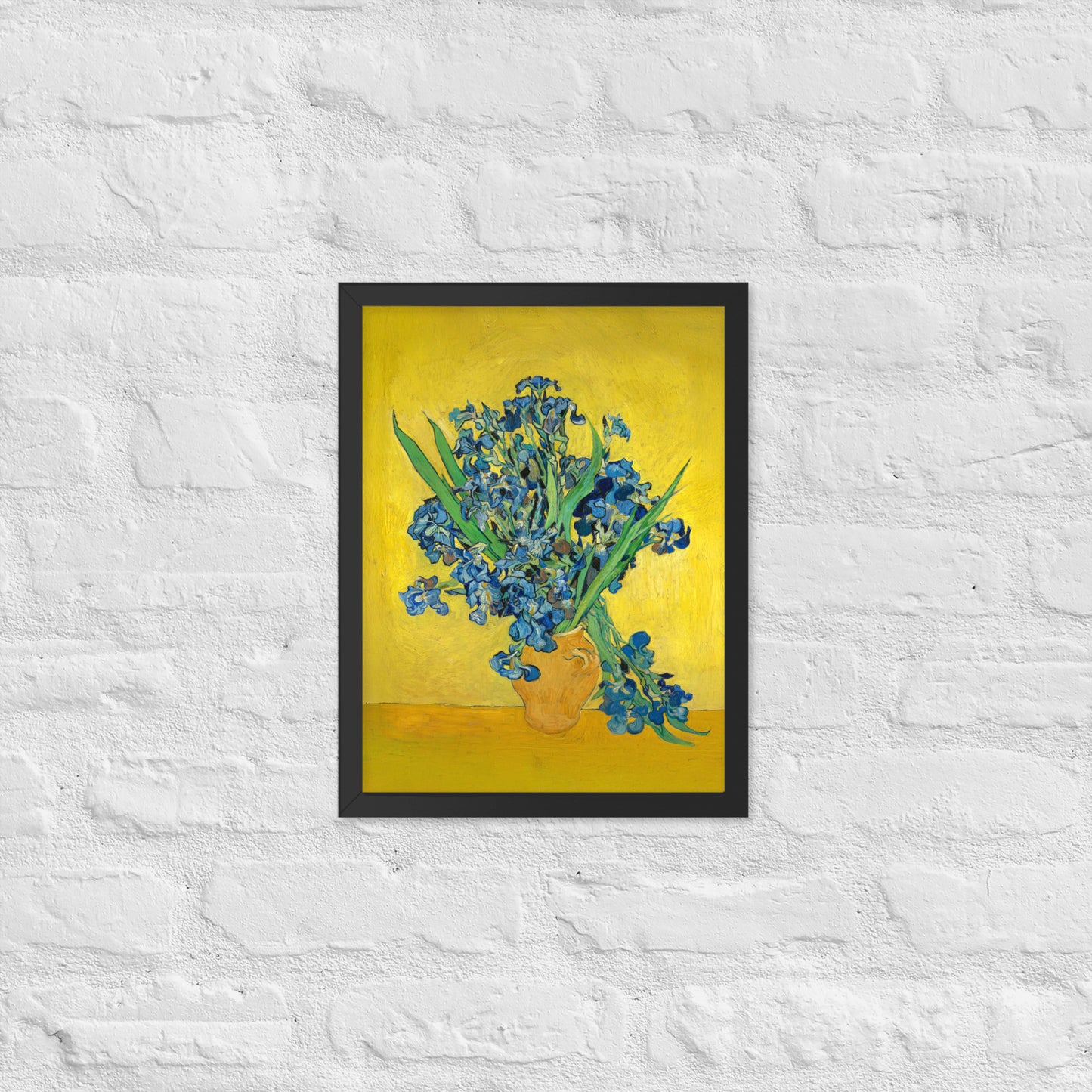Van Gogh's 'Vase with Irises Against a Yellow Background' on a 12"x16" black framed poster print on a white brick wall.