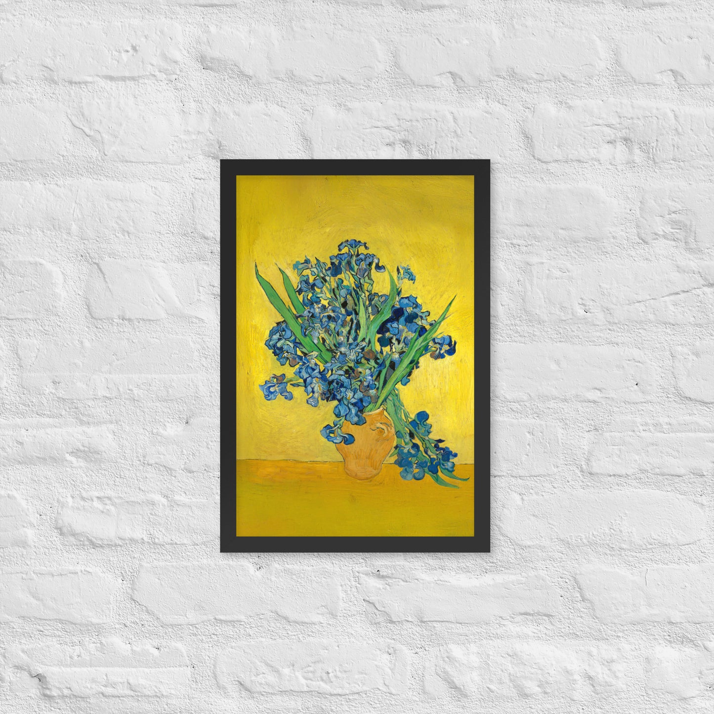Van Gogh's 'Vase with Irises Against a Yellow Background' on a 12"x18" black framed poster print on a white brick wall.