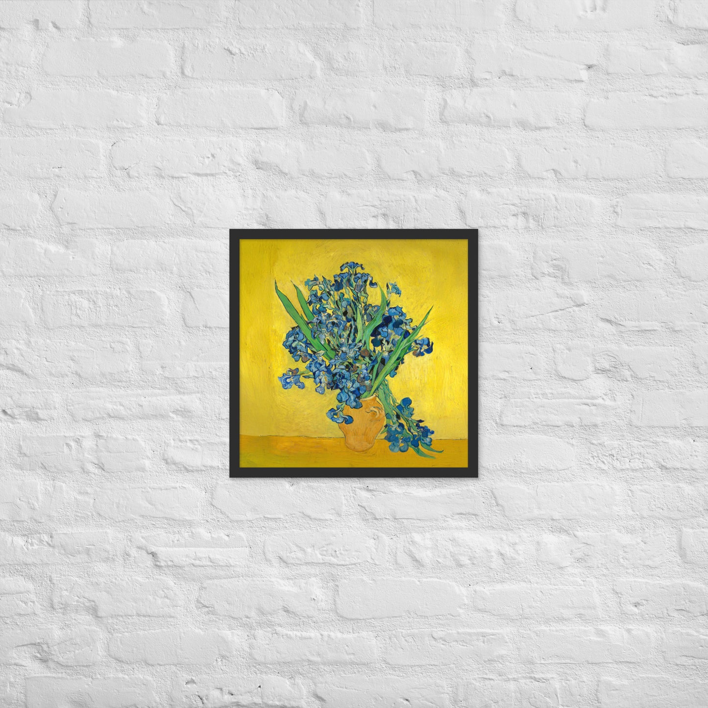 Van Gogh's 'Vase with Irises Against a Yellow Background' on a 18"x18" black framed poster print on a white brick wall.