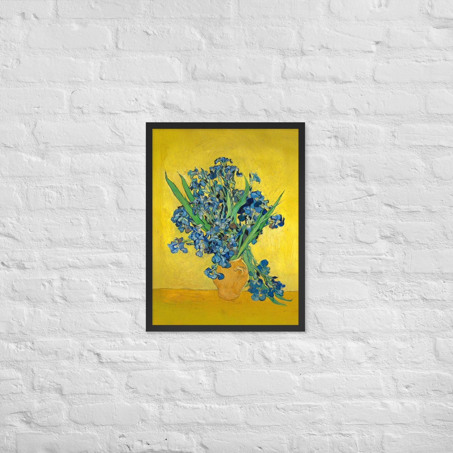 Van Gogh's 'Vase with Irises Against a Yellow Background' on a 18"x24" black framed poster print on a white brick wall.