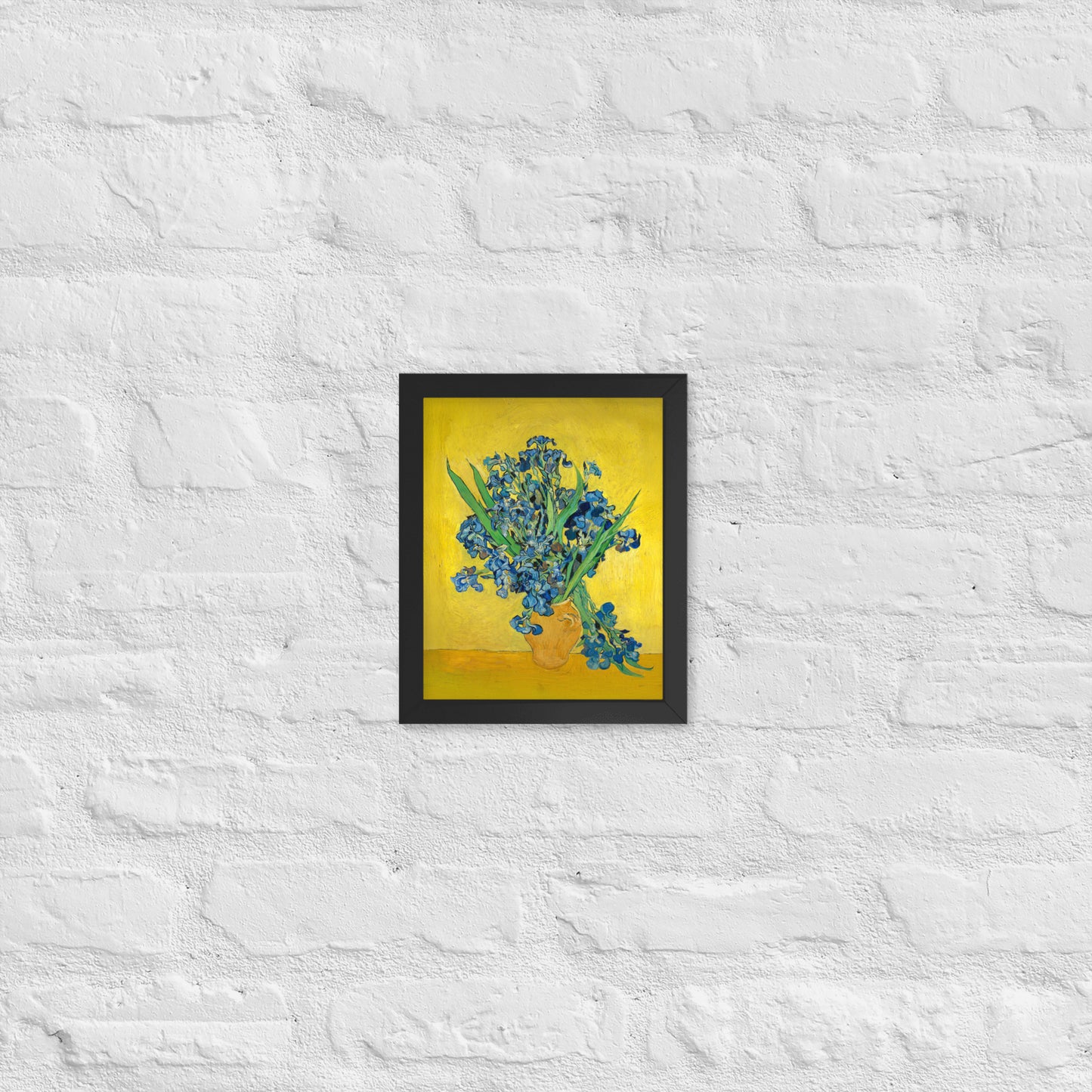 Van Gogh's 'Vase with Irises Against a Yellow Background' on a 8"x10" black framed poster print on a white brick wall.