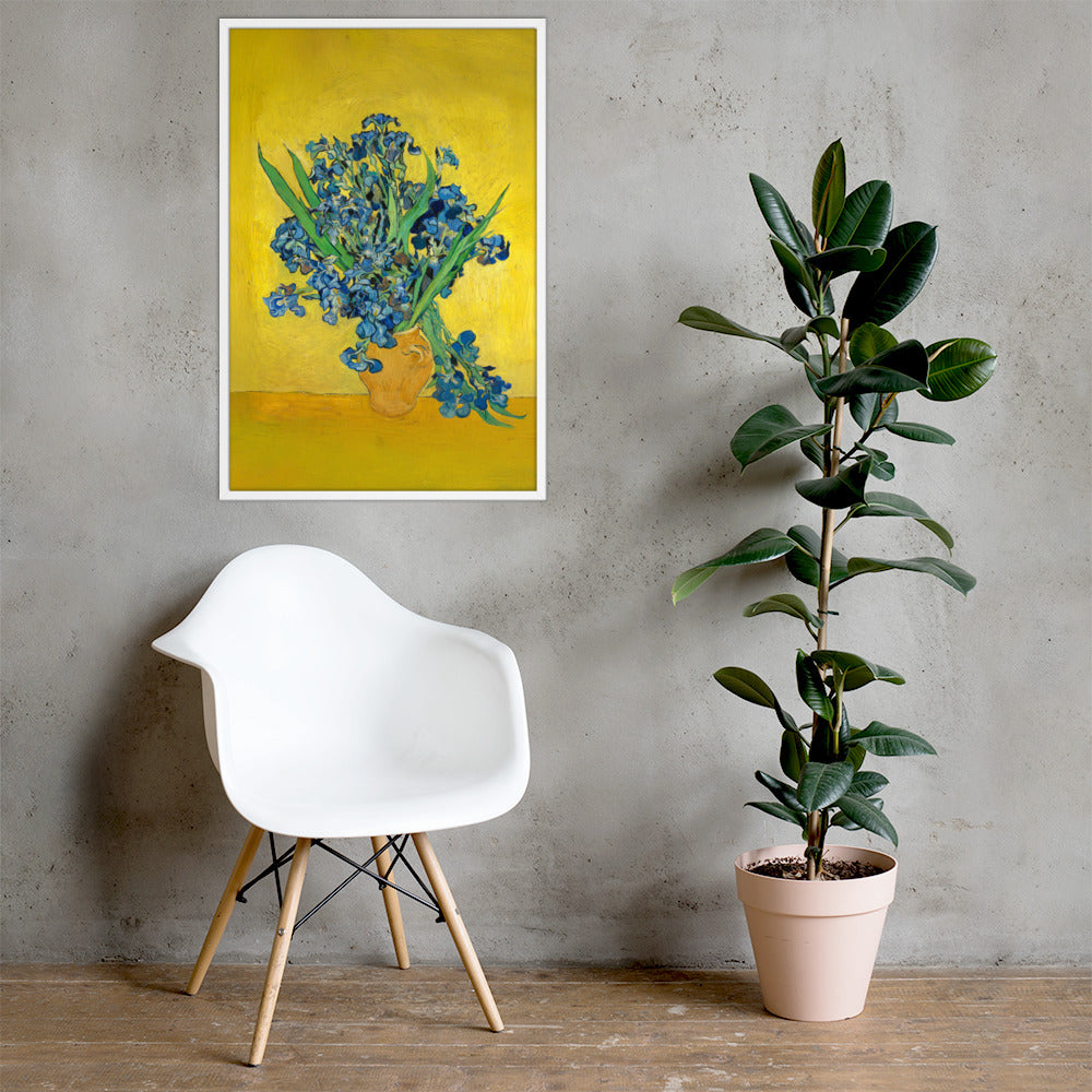 Van Gogh's 'Vase with Irises Against a Yellow Background' on a 24"x36" white framed poster print on a light gray concrete wall with a white chair and plant below.