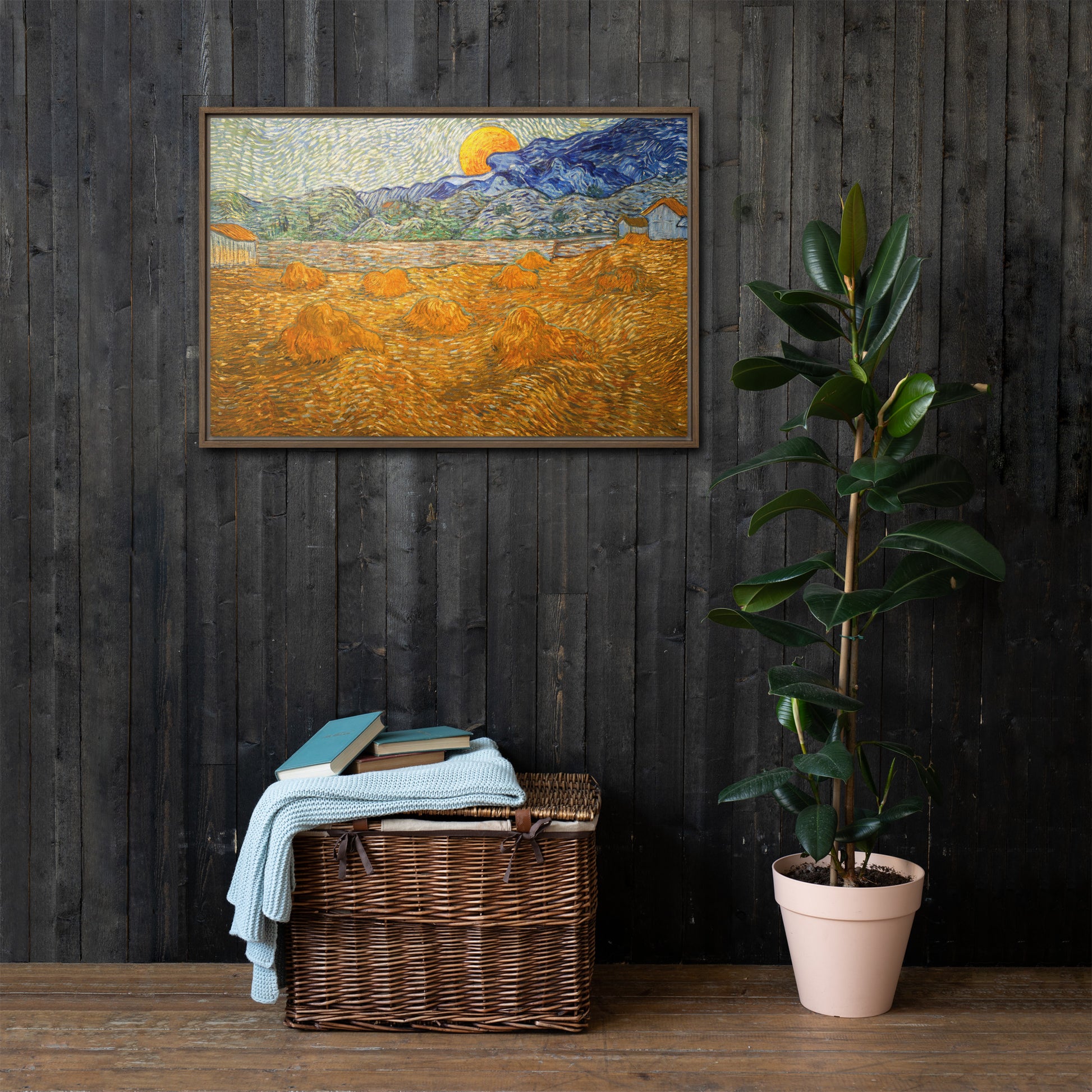 Van Gogh's 'Landscape with Wheat Sheaves and Rising Moon' on a 24"x36" brown framed canvas print on a dark wooden wall with a wicker bin and plant below.