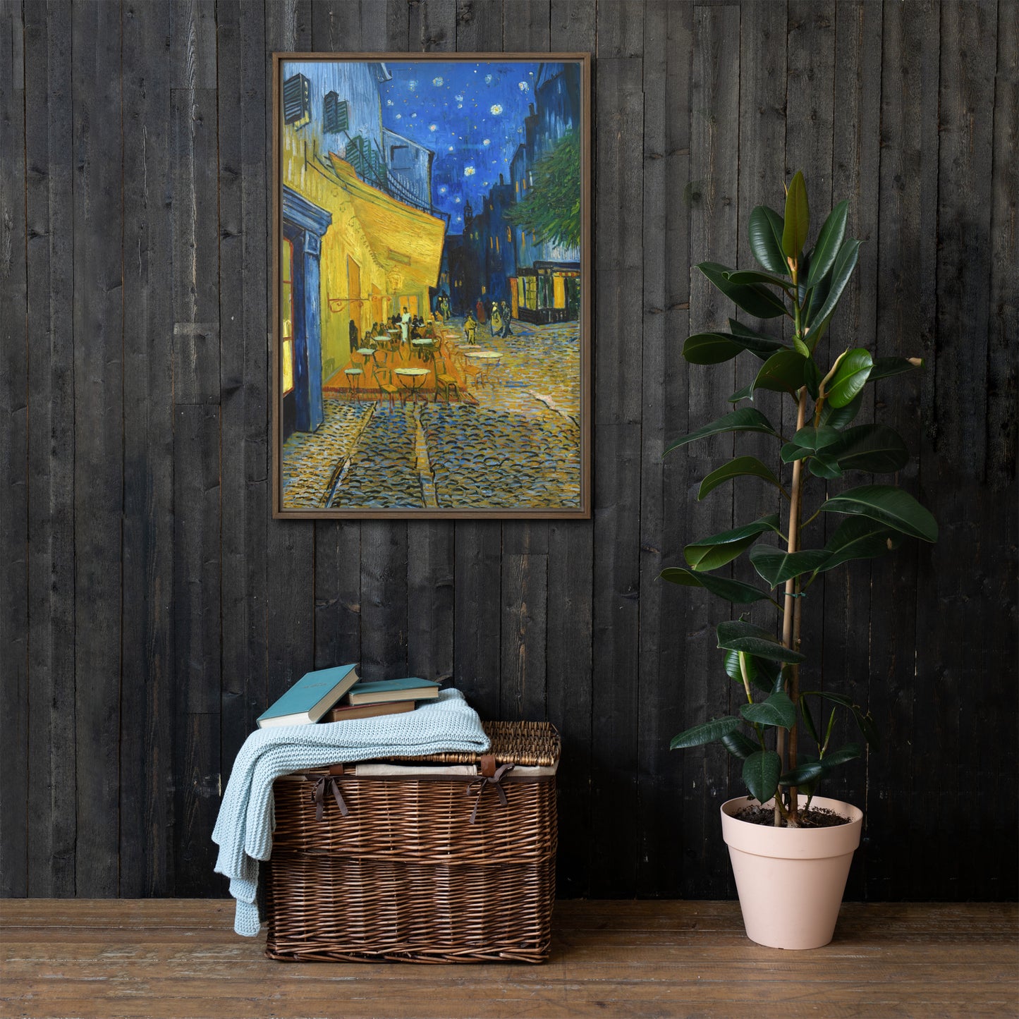 Van Gogh's 'Café Terrace at Night' on a 24"x36" brown framed canvas print on a dark wooden wall with a wicker bin and plant below.