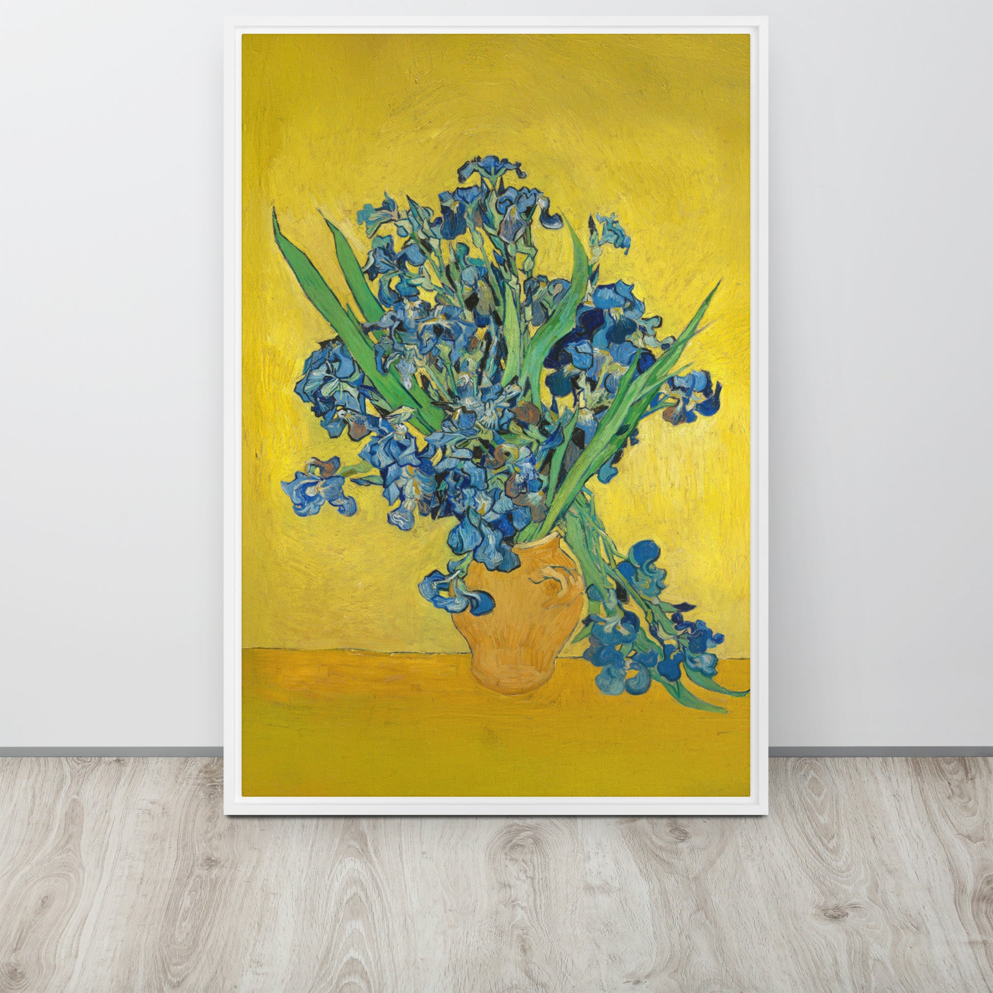 Van Gogh's 'Vase with Irises Against a Yellow Background' on a 24"x36" white framed canvas print sitting on a wooden floor leaning against a light gray wall.